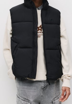 Personne portant un gilet sans manches noir matelassé par-dessus un pull crème et un jean en denim gris clair avec des poches.