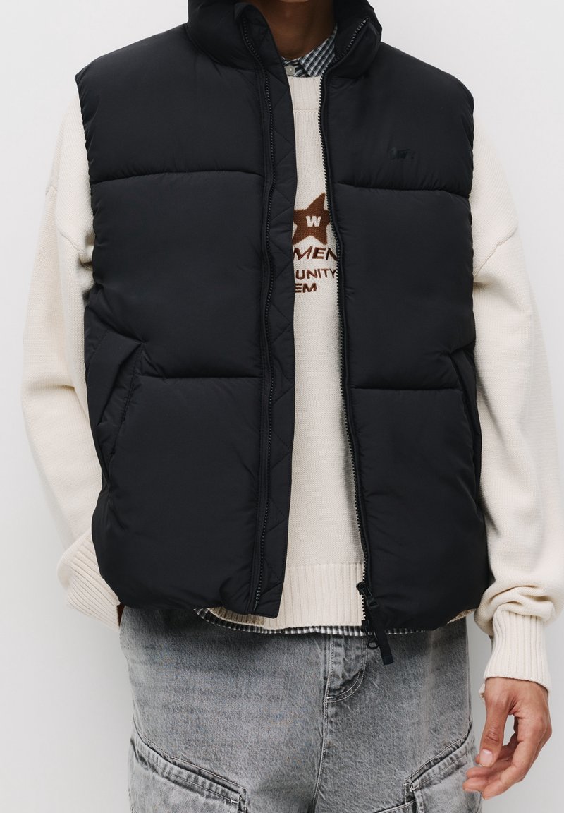 Personne portant un gilet sans manches noir matelassé par-dessus un pull crème et un jean en denim gris clair avec des poches.
