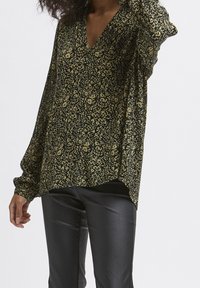 Blusa nera e verde con motivo floreale, scollo a V, maniche lunghe e vestibilità comoda, indossata sopra pantaloni neri attillati.