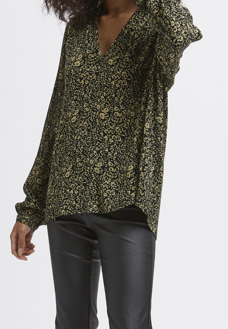 Blusa nera e verde con motivo floreale, scollo a V, maniche lunghe e vestibilità comoda, indossata sopra pantaloni neri attillati.