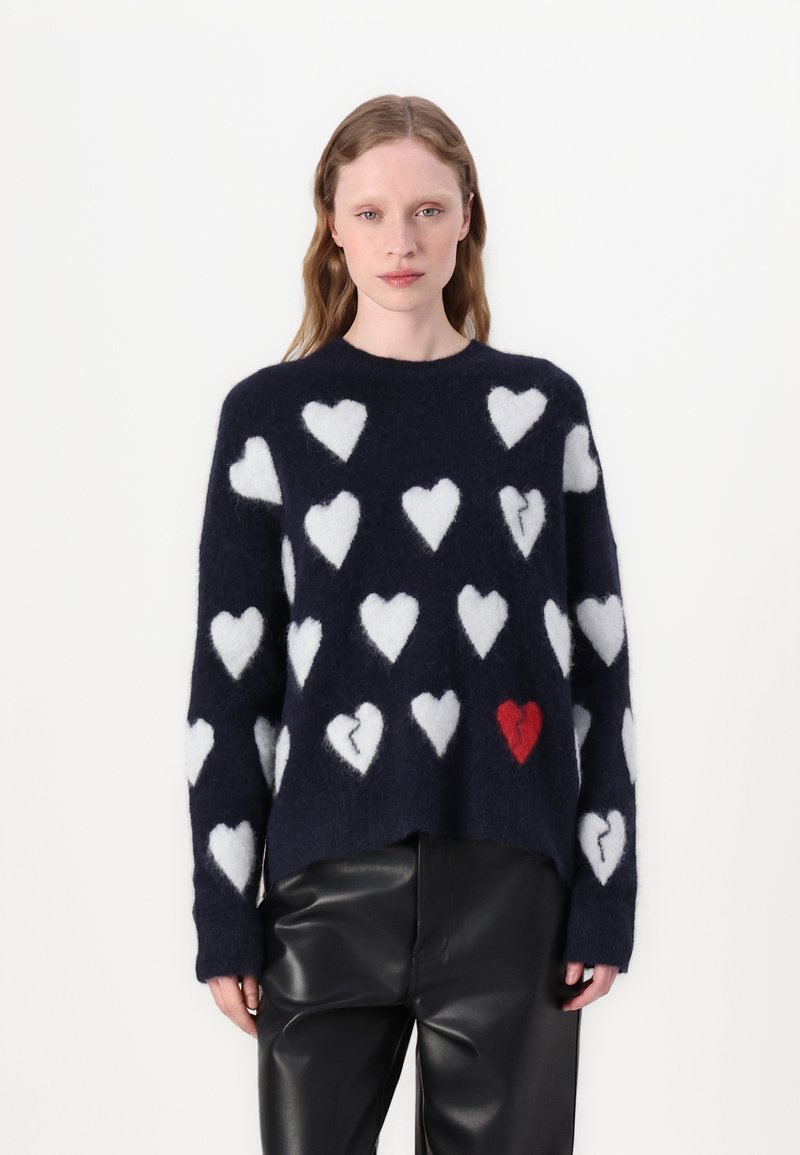 Maglione blu navy con motivo di cuori bianchi e uno rosso. Tessuto morbido e soffice, con vestibilità comoda e scollo rotondo. Indossati pantaloni neri.