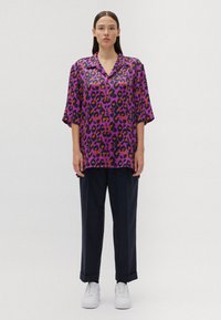Camicia viola a maniche corte con stampa leopardata nera e arancione, abbinata a pantaloni scuri e sneakers bianche. Tessuto morbido, vestibilità ampia.