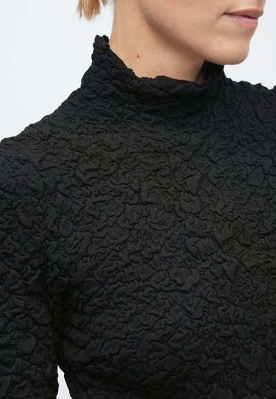 Naine, kes kannab tekstuuriga musta kõrge kaelusega kampsunit, mis on kärbitud, et näidata kaela ja alalist nägu ühevärvilise tausta vastu.