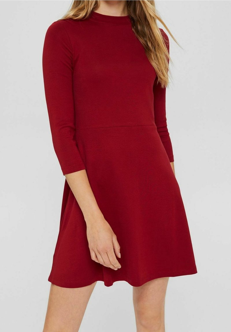 Punainen, polvipituinen mekko sileästä kankaasta, jossa on korkea kaulus ja kolmen neljänneksen hihat. Mekossa on istuva yläosa ja levenevä hame.