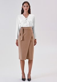 Donna che indossa un blouse bianco con lacci, una gonna avvolgente beige con fiocco laterale, tacchi neri, in piedi contro uno sfondo chiaro e semplice.