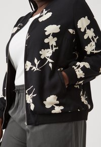 Svart bomberjacka med vitt blommönster, utrustad med dragkedja, ribbade muddar och avslappnad passform. Kombinerad med en vit topp och grå byxor.