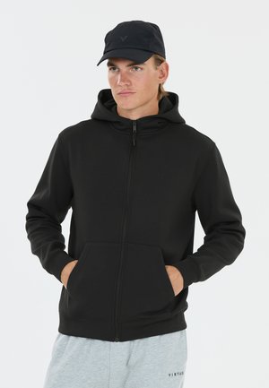 Young man wearing a black zip-up hoodie with hands in pockets, a black cap, and light gray sweatpants with "VIRTUS" text.