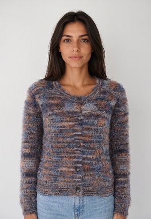 Vrouw met lang donker haar, gekleed in een zachte meerkleurige vest met knopen en lichtblauwe spijkerbroek, staand tegen een effen achtergrond.