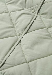 Lys grøn quiltet tæppe med en blød tekstur, prydet med knapdetaljer og diagonale syninger i et kvadratisk mønster.