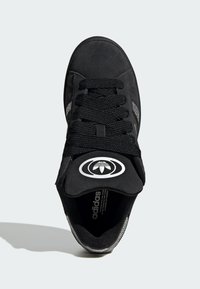 Crna sportska cipela s gornjim dijelom od brušene kože, teksturiranim vezicama i bijelim okruglim logotipom na jeziku, sa srebrnim detaljima sa strane.