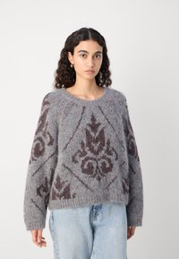 Grauer Pullover mit einer strukturierten, flauschigen Oberfläche, die ein braunes geometrisches Muster aufweist. Rippenkragen und - manschetten, lockere Passform, kombiniert mit hellblauen Jeans.