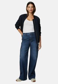 Navy bomber jacket, white tank top, wide-leg dark denim cargo pants with front pockets, and light-colored shoes. Smooth fabric and relaxed fit.