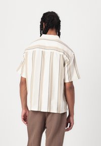 Chemise à manches courtes avec une base blanche ornée de rayures verticales beiges et noires, coupe ample, col et tissu texturé.