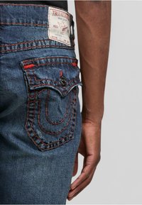 Tmavomodré džínsy z denimu s výrazným šitím v tvare "U" na zadných vreckách, červenými akcentmi a s označením značky na páse.