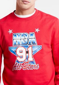 Homme portant un sweat-shirt rouge avec le texte bleu et blanc "NBA 91 All-Stars" et des motifs d'étoiles sur la poitrine, par-dessus un t-shirt à col rond blanc.