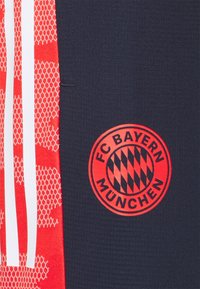 Námořnické sportovní šortky s červenými síťovými vzory a bílými pruhy. Uprostřed logo: kruhový design s textem "FC Bayern München" a diamantovým vzorem.