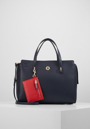 Navy blue handbag with gold logo and hardware, featuring double handles and a detachable red zippered pouch hanging on the front.