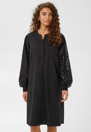 Mujer con cabello rizado que lleva un vestido negro hasta la rodilla con mangas largas de lentejuelas y un escote abotonado, de pie frente a un fondo liso.