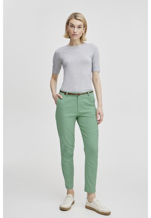 Kvinde stående iført en tætsiddende lys grå kortærmet top, mintgrønne bukser med brunt bælte og hvide sneakers mod en ensfarvet baggrund.