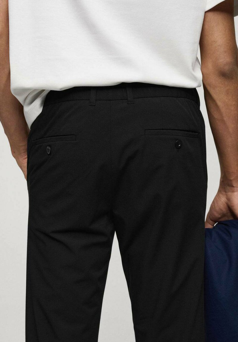 Un pantalon noir avec des lignes épurées, deux poches arrière avec des boutons et une texture de tissu lisse. Une main tient un objet bleu marine à côté de la taille.