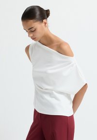 Top bianco, off-shoulder, realizzato in tessuto morbido con linee drappeggiate, maniche corte e vestibilità comoda, abbinato a pantaloni bordeaux. Texture liscia e design elegante.