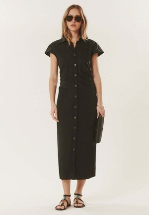 Femme portant une robe midi noire à boutons, des sandales noires, tenant une pochette noire texturée et portant des lunettes de soleil foncées, debout devant un fond uni.