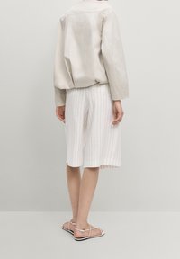 Personen bærer en lys beige langærmet top, hvide pinstripede knælange shorts og hvide flade sandaler med stropper, og står foran en ensfarvet væg.
