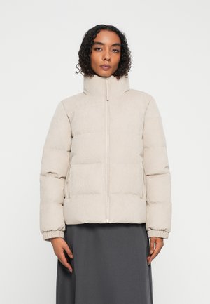 Frau mit lockigem Haar, die eine beige Steppjacke trägt, die bis oben zugezippt ist, und vor einem schlichten weißen Hintergrund steht, mit entspannten Händen an ihren Seiten.