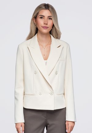 Femme aux longs cheveux blonds portant un blazer blanc à double boutonnage, des boucles d’oreilles en or, un collier et un pantalon gris sur un fond uni.