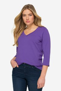 Lila langärmliges Oberteil mit V-Ausschnitt, aus weichem Material, kombiniert mit dunkelblauen Jeans. Der Stoff wirkt glatt und leicht.