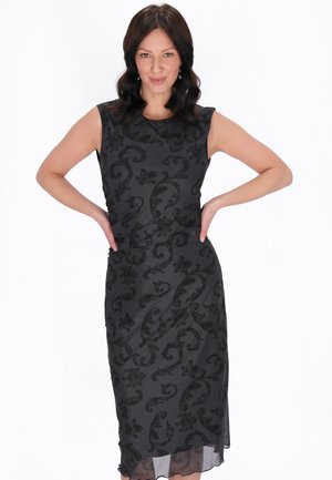 Femeie cu păr închis la culoare purtând o rochie neagră fără mâneci cu modele florale, stând cu mâinile pe șolduri, pe un fundal alb.