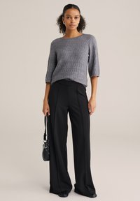 Grijze gebreide trui met een gestructureerd patroon, gecombineerd met zwarte, high-waisted broek met wijde pijpen. De outfit wordt aangevuld met een kleine zwarte handtas.