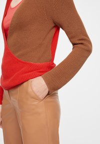 Cardigan en tricot présentant un design à fente avec des sections marron et rouge, associé à un pantalon en cuir beige ajusté, mettant en valeur le contraste de textures et de couleurs.