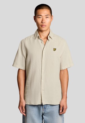 Junger Mann trägt ein beige-farbenes Kurzarm-Hemd mit Knöpfen und einem gelben Vogel-Logo auf der Brust sowie eine hellblaue Jeans, steht vor einem grauen Hintergrund.