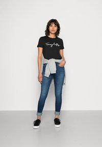 Melns T-krekls ar baltu "Tommy Hilfiger" logo, apvienots ar ziliem pieguļošiem džinsiem un pelēku pūkainu kreklu, kas sasiets ap vidukli. Melni apavi ar baltiem zoliem.