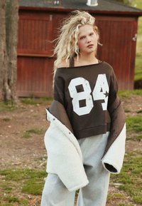 Felpa corta marrone con grafica bianca "84", abbinata a pantaloni tuta grigi e a una giacca oversize color crema foderata in shearling.