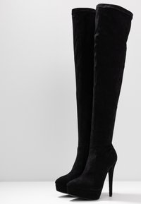Un par de botas de plataforma altas de ante negro con tacones stiletto altos, sobre un fondo blanco liso.