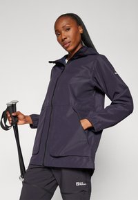 Giacca impermeabile blu navy con cappuccio, chiusura a zip e due grandi tasche. Presenta polsini elasticizzati e un materiale traspirante. Abbinata a pantaloni scuri.