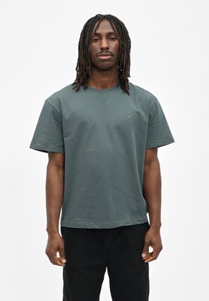 Man with shoulder-length dreadlocks wearing a green short-sleeve t-shirt and black pants standing against a plain white background.