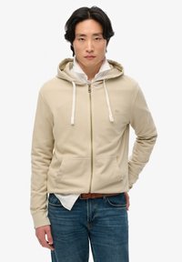 Sudadera con cremallera beige, con cintas de ajuste, un bolsillo frontal y un logo sutil. Se lleva sobre una camisa blanca con cuello y combinada con vaqueros azules.