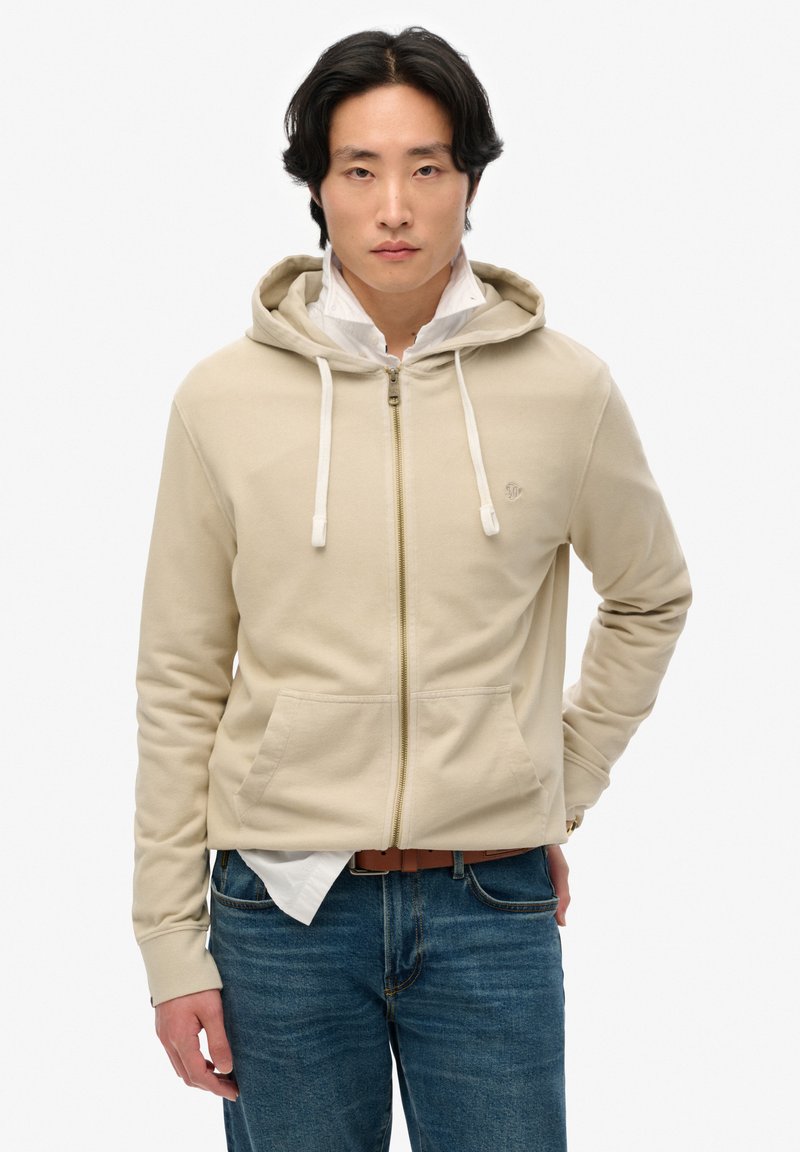 Sudadera con cremallera beige, con cintas de ajuste, un bolsillo frontal y un logo sutil. Se lleva sobre una camisa blanca con cuello y combinada con vaqueros azules.