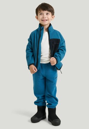Young boy smiling, wearing a blue fleece jacket and pants, white shirt, and black boots, standing against a plain light background.
