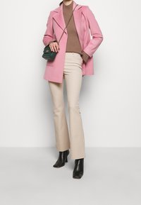 Manteau en laine rose, col roulé ajusté beige à côtes, pantalon flare beige, bottines noires à talons et sac bandoulière vert avec des accessoires en or.
