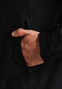 Schwarze Jacke mit Nahaufnahme einer Hand, die in eine Reißverschlusstasche greift; zeichnet sich durch glatte Textur und strapazierfähigen Stoff aus.