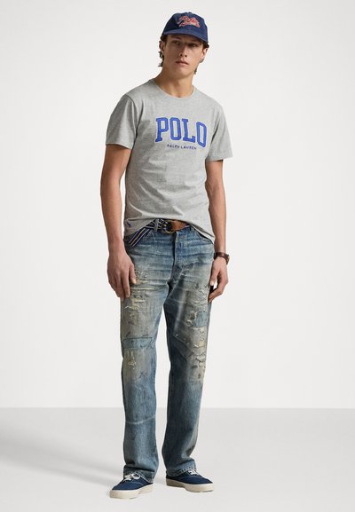 Tricou gri din bumbac cu text albastru "POLO" și "Ralph Lauren" dedesubt. Blugi de denim purtați cu petice distruse. Adidași albaștri. Capac bleumarin.