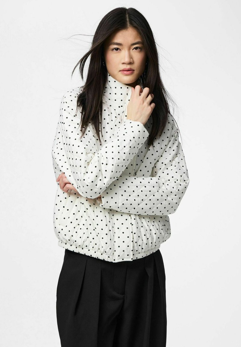 Jeune femme aux longs cheveux noirs portant une veste matelassée blanche à pois noirs et un pantalon noir à jambes larges sur fond blanc.