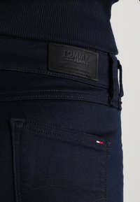 Mörk denimjeans i marinblått med en ribbad midja och en svart läderlapp märkt "TOMMY JEANS", som har en kontrasterande liten accent i röd, vit och blå.