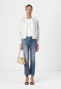 Weiße gemusterte Jacke, weißes T-Shirt, blaue Jeans, schwarze High-Heelsandalen und eine beigefarbene Handtasche. Modell steht vor einem schlichten Hintergrund.