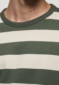 Nahaufnahme des Nackens und des oberen Brustbereichs, der ein grün-weiß gestreiftes Rundhalsausschnitt-Shirt trägt, mit sichtbaren Hautmolen.