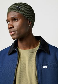Gorro de canalé en verde olivo con un parche de logo negro, usado con una chaqueta azul que tiene un cuello de pana negro y una camisa verde claro debajo.